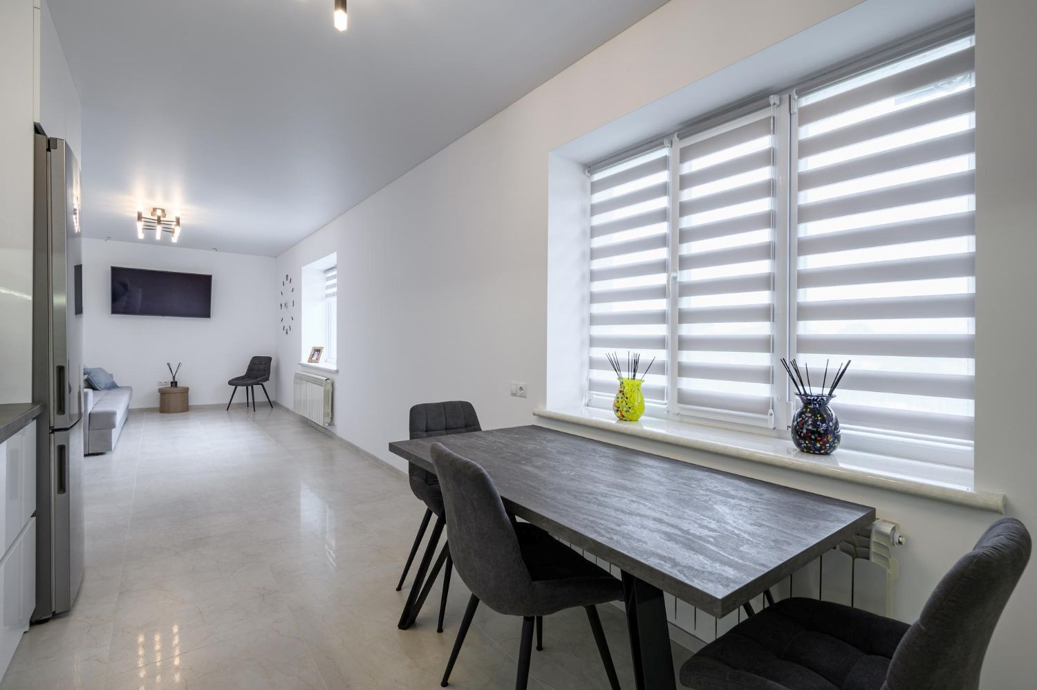 Modern zebra roller blinds installed on a window in a minimalist white dining room with a dark wooden table.
