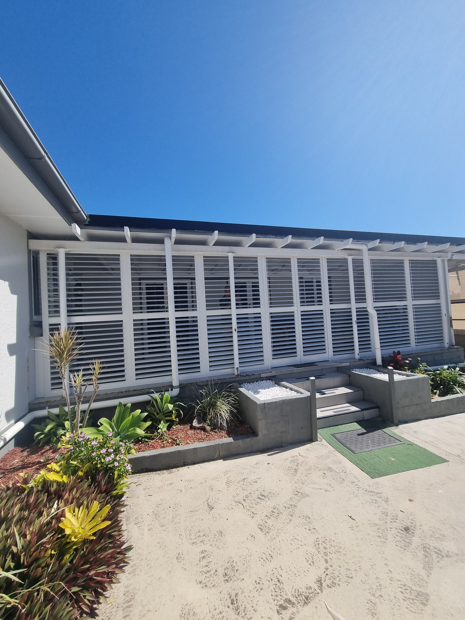 Modern home exterior with white louvered shutters, landscaped garden beds, and a clear blue sky.