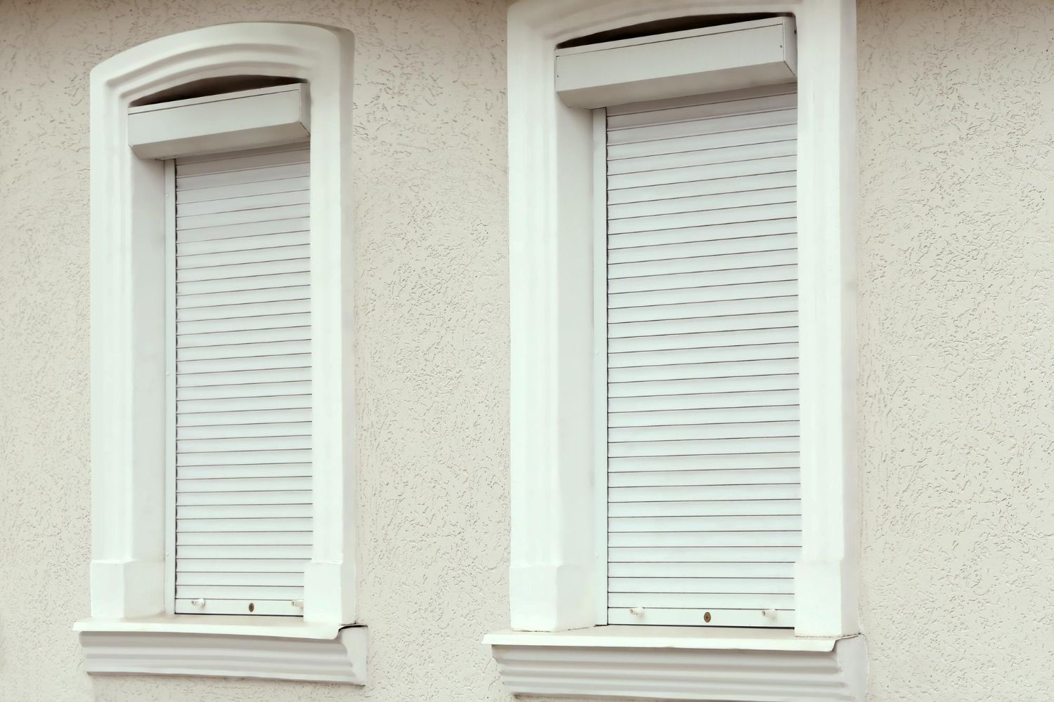 Exterior windows with white roller shutters on a stucco wall.